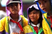 Three curious youngsters wearing colorful rain gear over white
shirts and red scarfs, the uniform typically worn by Vietnamese
school children, stop on their way home to try out their English
with two Americans having coffee at a sidewalk cafe. 90% of the
Vietnamese population can read.