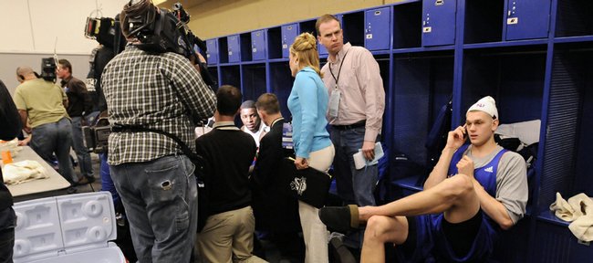 Kansas center Cole Aldrich talks on his cell phone in a pair of house slippers as his teammate Sherron Collins is surrounded by media members in the Jayhawks' locker room Thursday, March 26, 2009 at Lucas Oil Stadium in Indianapolis.